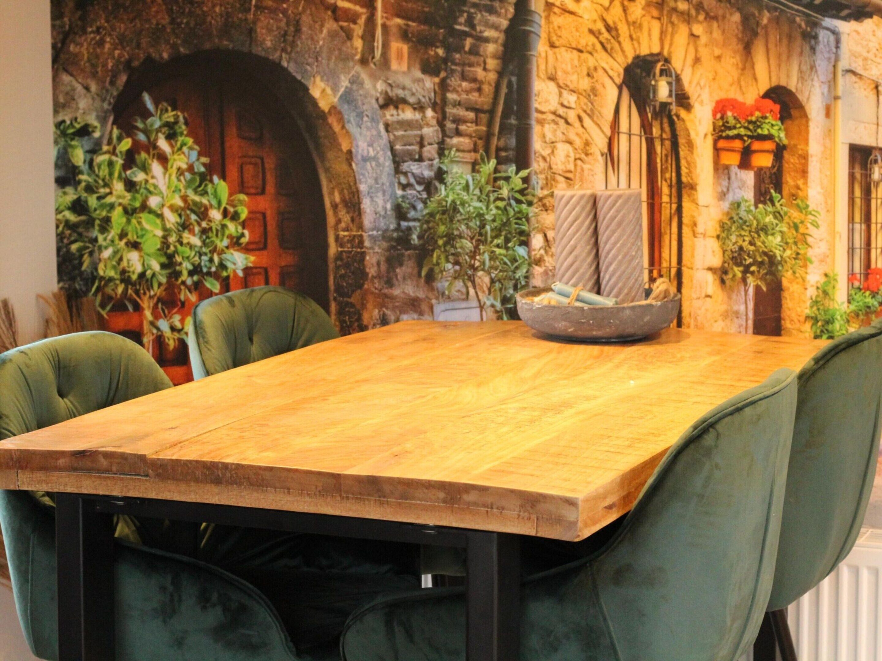 Een houten eettafel met groen gestoffeerde stoelen, geplaatst tegen een stenen muur met een houten deur en potplanten.