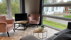 A modern living room with a large window, a flat-screen TV, and two pink armchairs. - Charming Chalet With Spacious Enclosed Garden (Hippolytushoef)