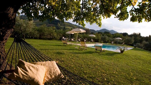 A hammock with a cushion, a pool, lounge chairs, and umbrellas in a grassy outdoor area with trees and hills in the background. - Spacious Holiday Home in Brisighella (Brisighella)