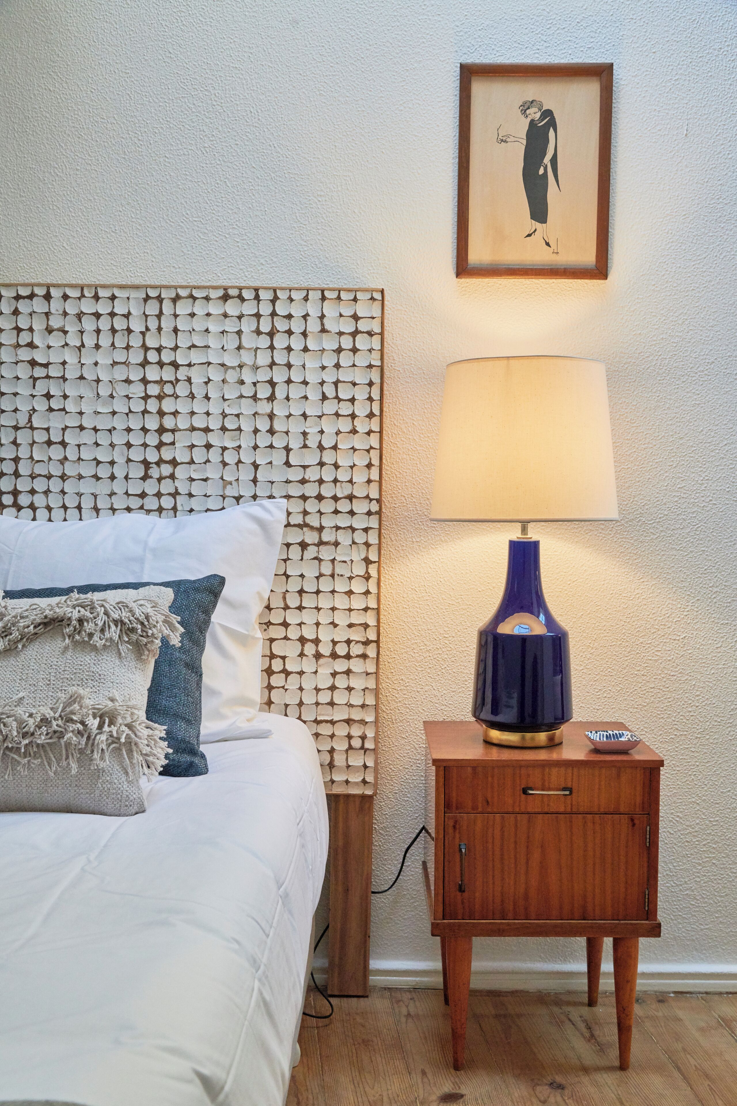 A bed with white bedding, a headboard with a textured pattern, a bedside lamp, a small wooden nightstand, and a framed picture on the wall.