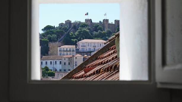 A view through a window of a building with a castle-like structure on a hill, a flag, and a tiled roof. - Central, Comfy And Light-Filled Apt, By Timecooler (Lisbon)
