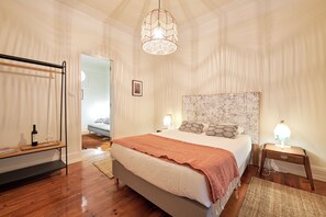 A bedroom with a bed, bedside lamps, a hanging light fixture, a wooden floor, and a door leading to another room.