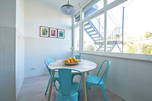 Un comedor con una mesa redonda, cuatro sillas color turquesa, un cuenco de limones y una ventana que da a una escalera.