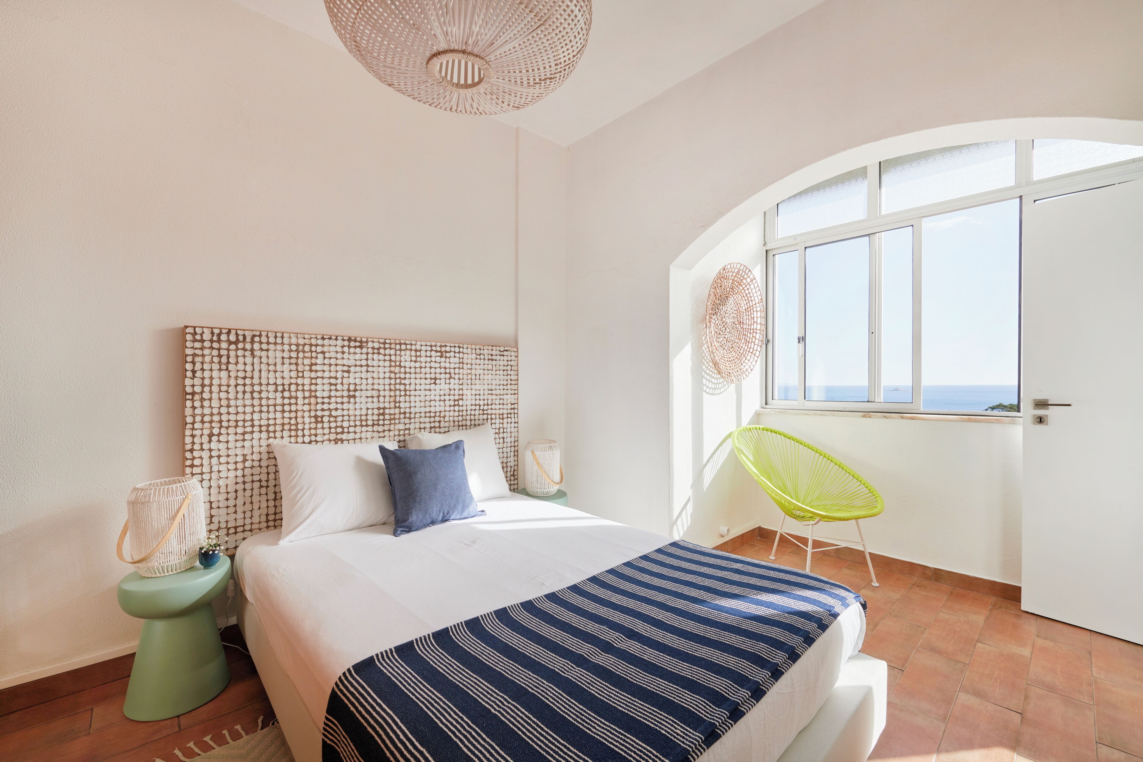 A bedroom with a bed, a bedside table, a chair, a window, and a ceiling light.