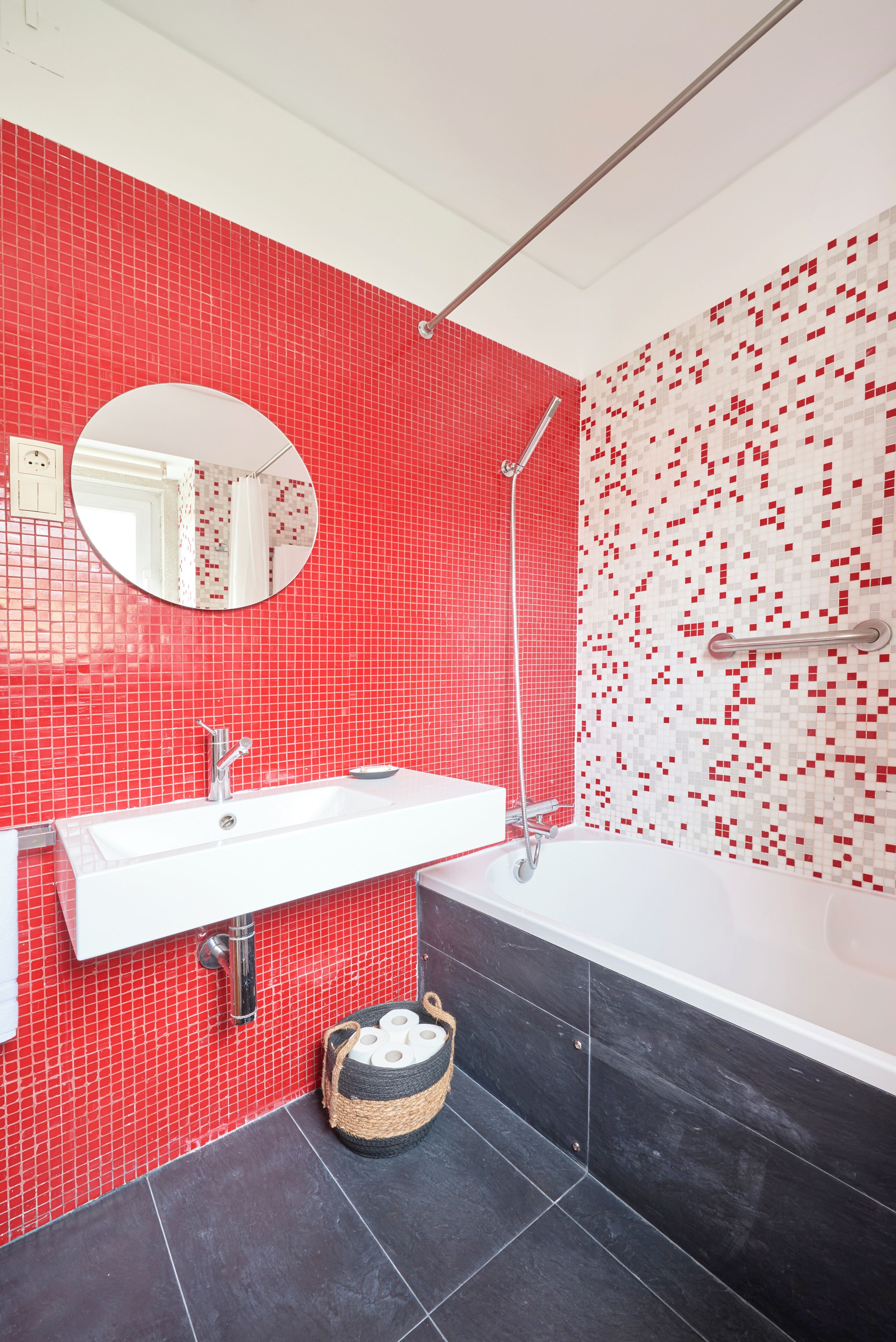 Un baño con pared de azulejos rojos, lavamanos blanco, espejo, ducha y suelo de baldosas negras.