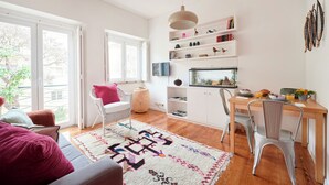 Una sala de estar luminosa con un sofá, una mesa de comedor con sillas, una estantería y un ventanal grande.