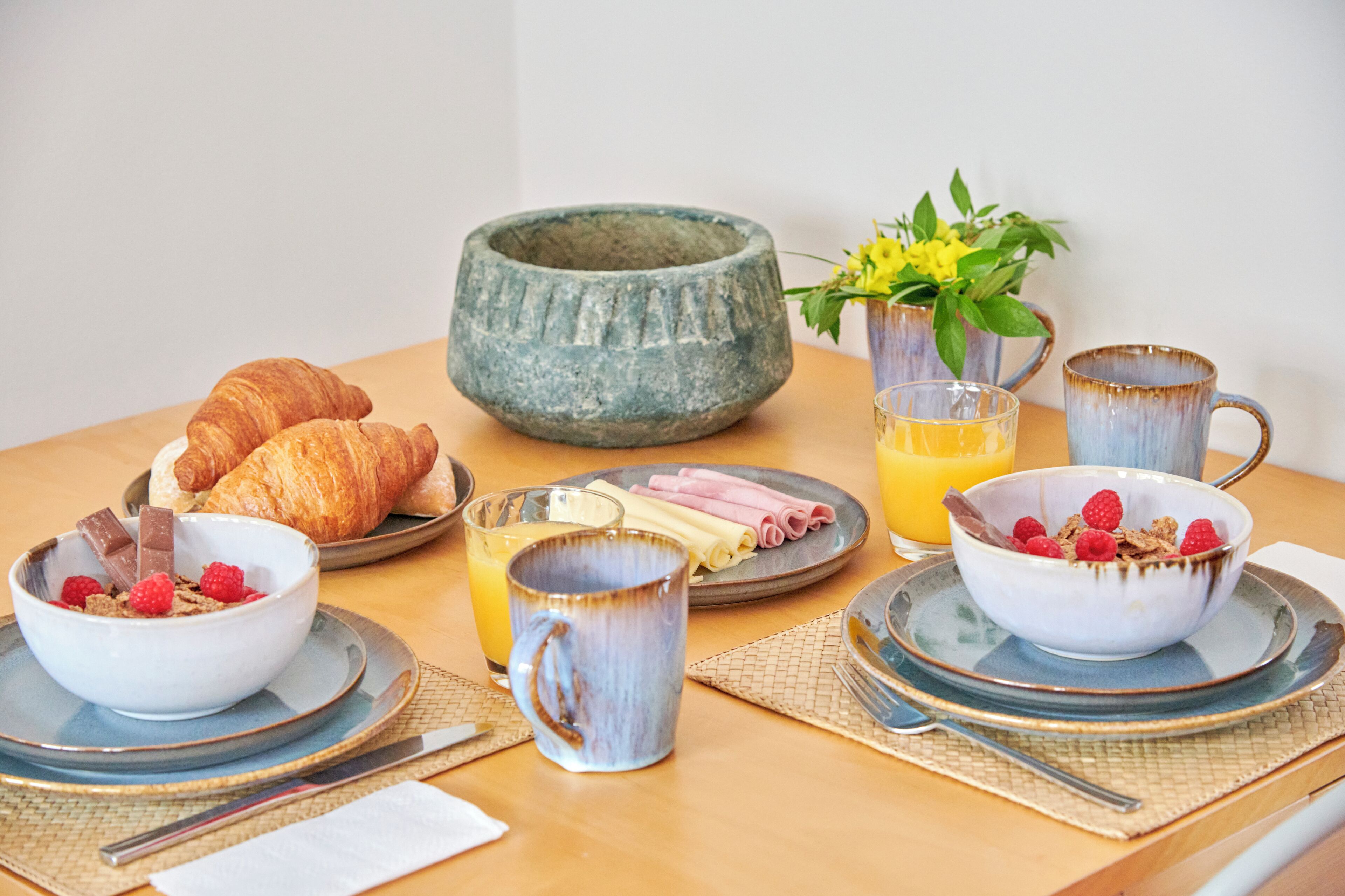 Ein Frühstückstisch mit Croissants, einer Schüssel Müsli mit Himbeeren, einem Glas Orangensaft und einer Keramiktasse.