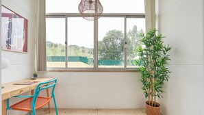 A room with a wooden desk, a blue chair, a woven mat, a potted bamboo plant, and a large window with a view of greenery. - Light Filled & Renovated Apartment, By Timecooler (Lisbon)