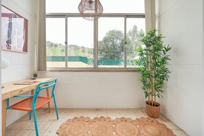 Une pièce avec un bureau en bois, une chaise bleue, un tapis tissé, un pot de bambou et une grande fenêtre donnant sur un paysage verdoyant.
