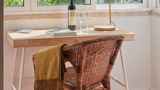 Ein Zimmer mit Schreibtisch, Korbsessel, einer Weinflasche, Weingläsern und einer Lampe.