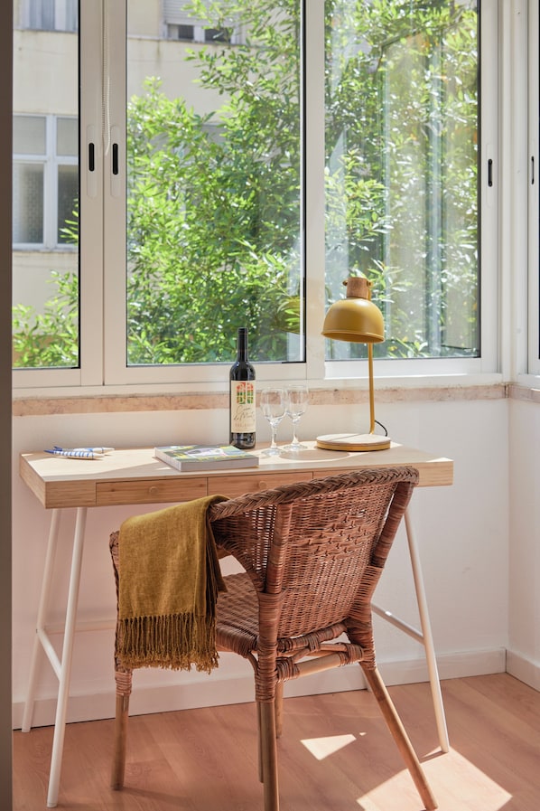 A room with a desk, wicker chair, a bottle of wine, wine glasses, and a lamp.