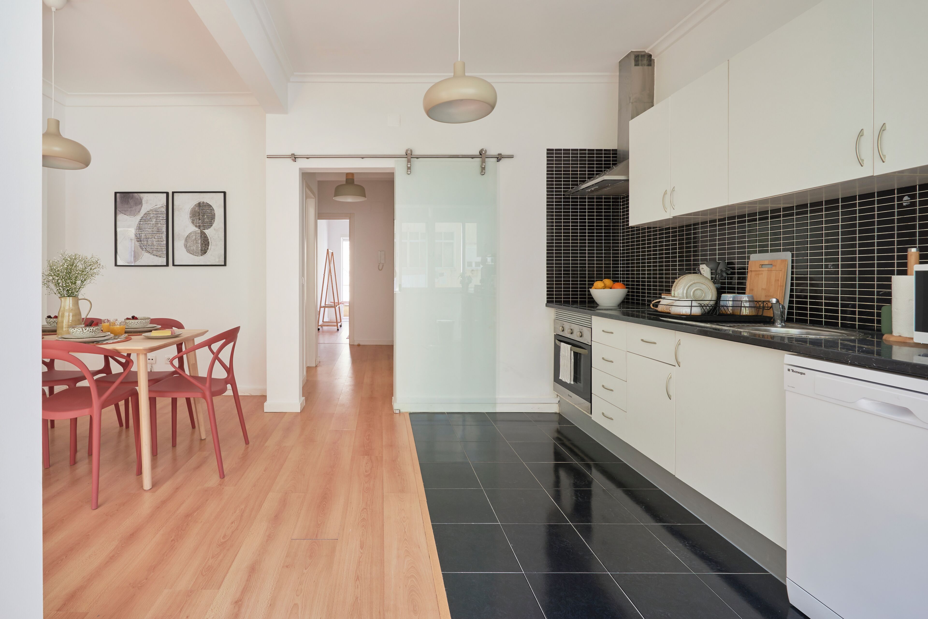 Una cocina moderna con armarios blancos, baldosas negras y una mesa de comedor de madera con sillas rojas.