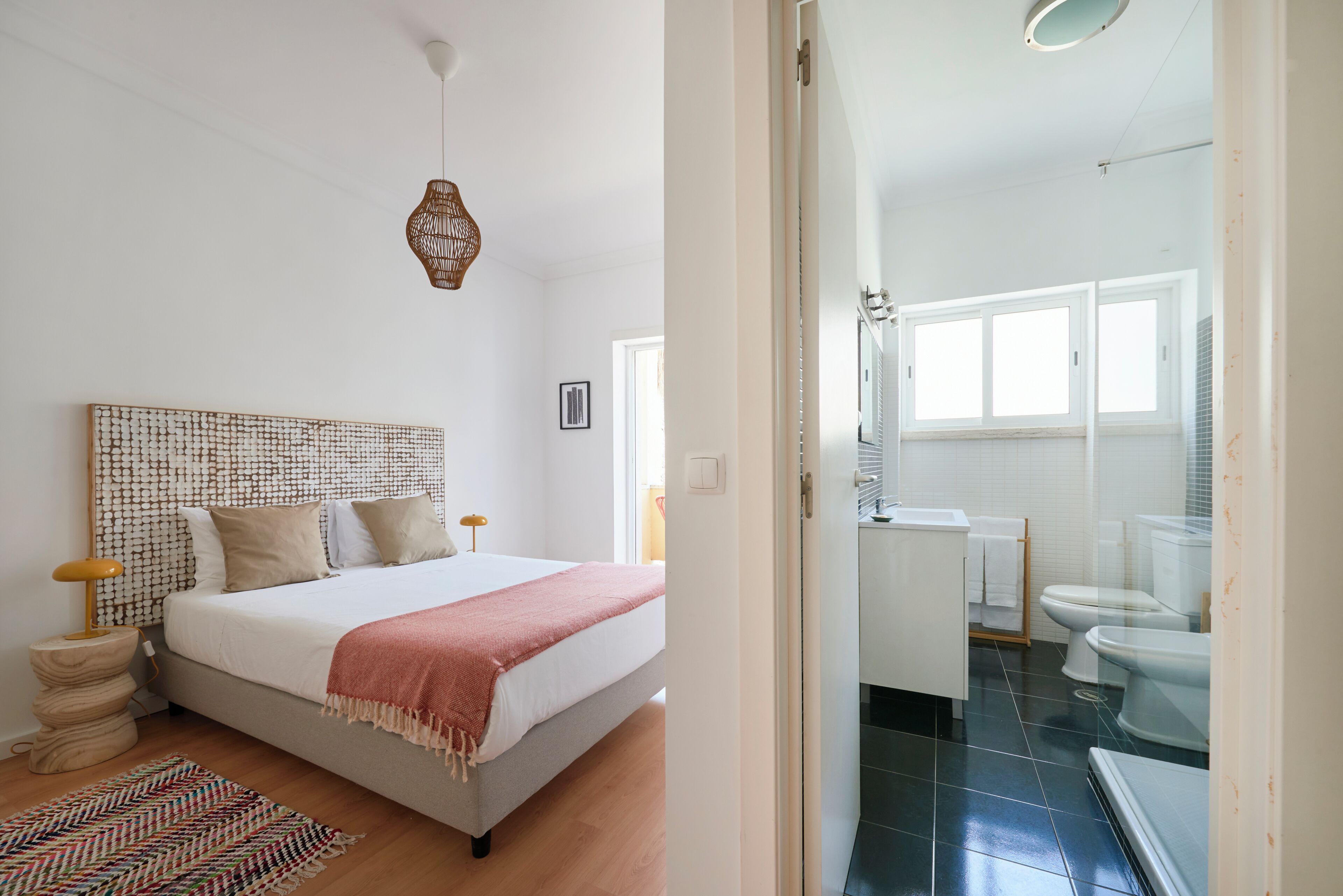 Un dormitorio con cama, mesitas de noche, cabecero tejido, lámpara colgante y vista al baño.