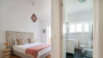 Un dormitorio con cama, mesitas de noche, cabecero tejido, lámpara colgante y vista al baño.