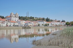 A waterfront view of a town with buildings, a river, and a clear sky. - Lightful Refuge On The River Front, By Timecooler (Alcacer do Sal)
