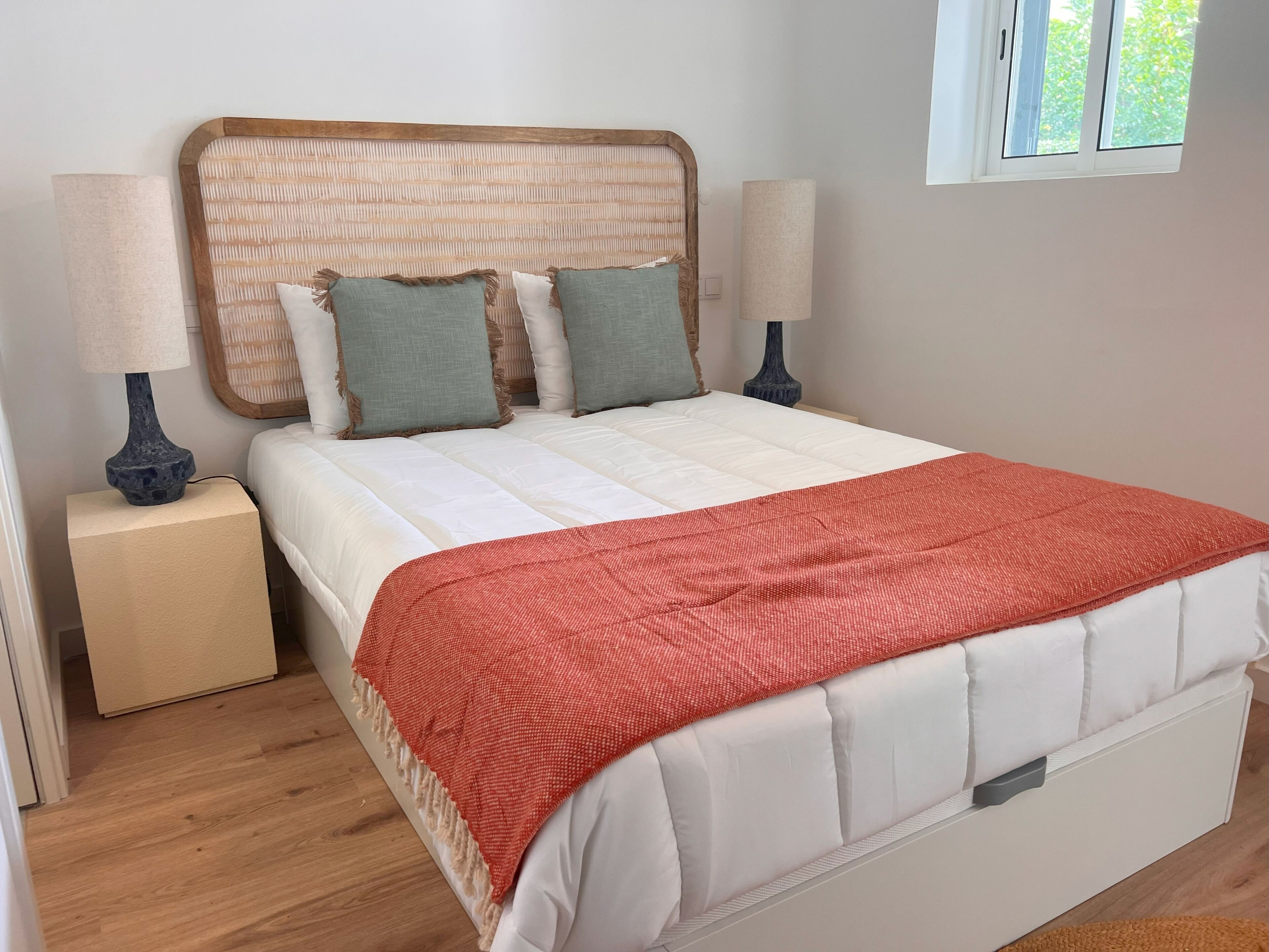 A neatly made bed with a wooden headboard, two bedside lamps, and a window with a view of greenery.