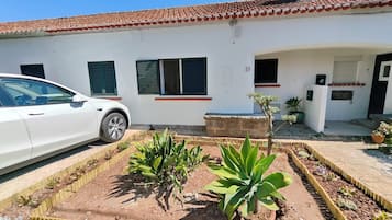 Un edificio blanco con techo de tejas rojas, un coche blanco estacionado adelante y un jardín con plantas y un pequeño árbol.