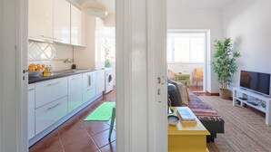 A modern kitchen with white cabinets, a washing machine, and a dining area with a sofa and TV. - Central & Sunny 2Br Apt Near Beach, By Timecooler (Setubal)