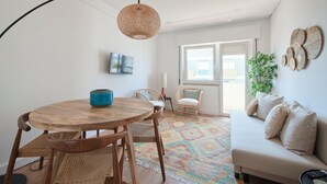 A modern living room with a round wooden table, wicker chairs, a beige sofa, a colorful rug, and a large window. - Sunset Bay View Renovated Apt, By Timecooler (Setubal)