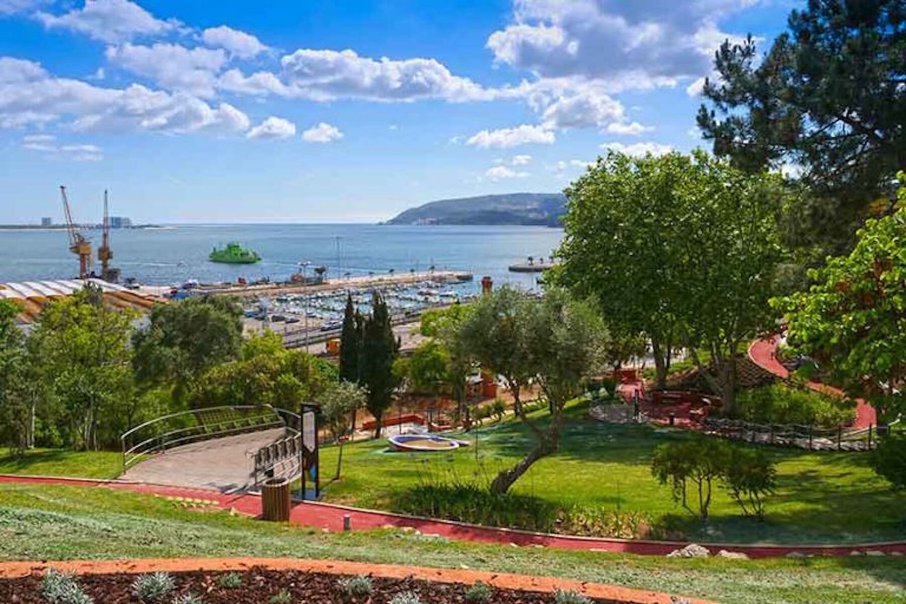 A coastal park with a playground, walking paths, and a view of the sea and distant buildings.
