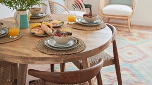 A round wooden dining table with placemats, a vase with flowers, and various breakfast items including croissants, fruit, and a bowl of cereal. - Sunset Bay View Renovated Apt, By Timecooler (Setubal)