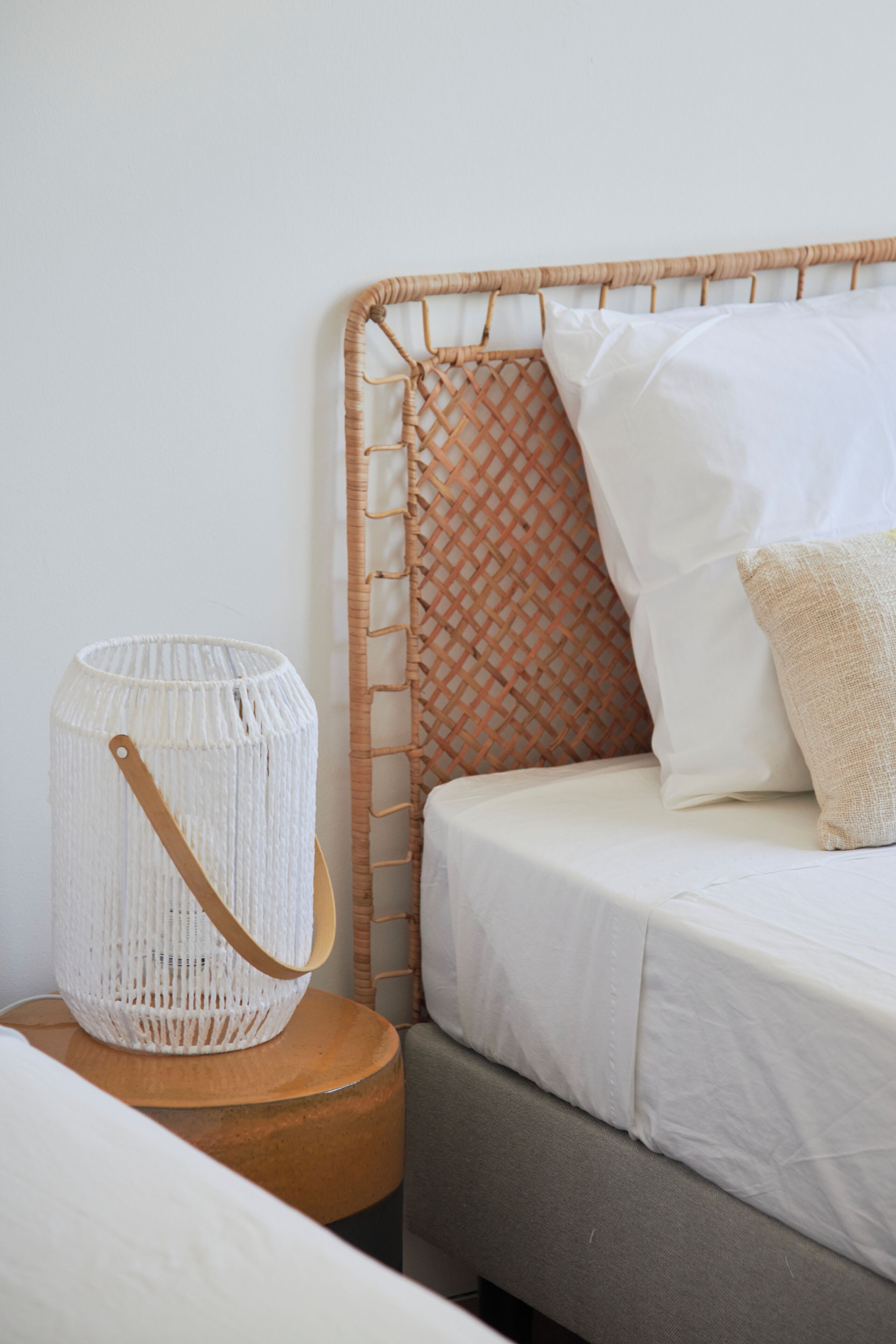 Una cama con un cabecero tejido, una almohada blanca, una almohada beige y una lámpara blanca de fibra sobre una mesita de noche de madera.