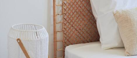 Una cama con un cabecero tejido, una almohada blanca, una almohada beige y una lámpara blanca de fibra sobre una mesita de noche de madera.