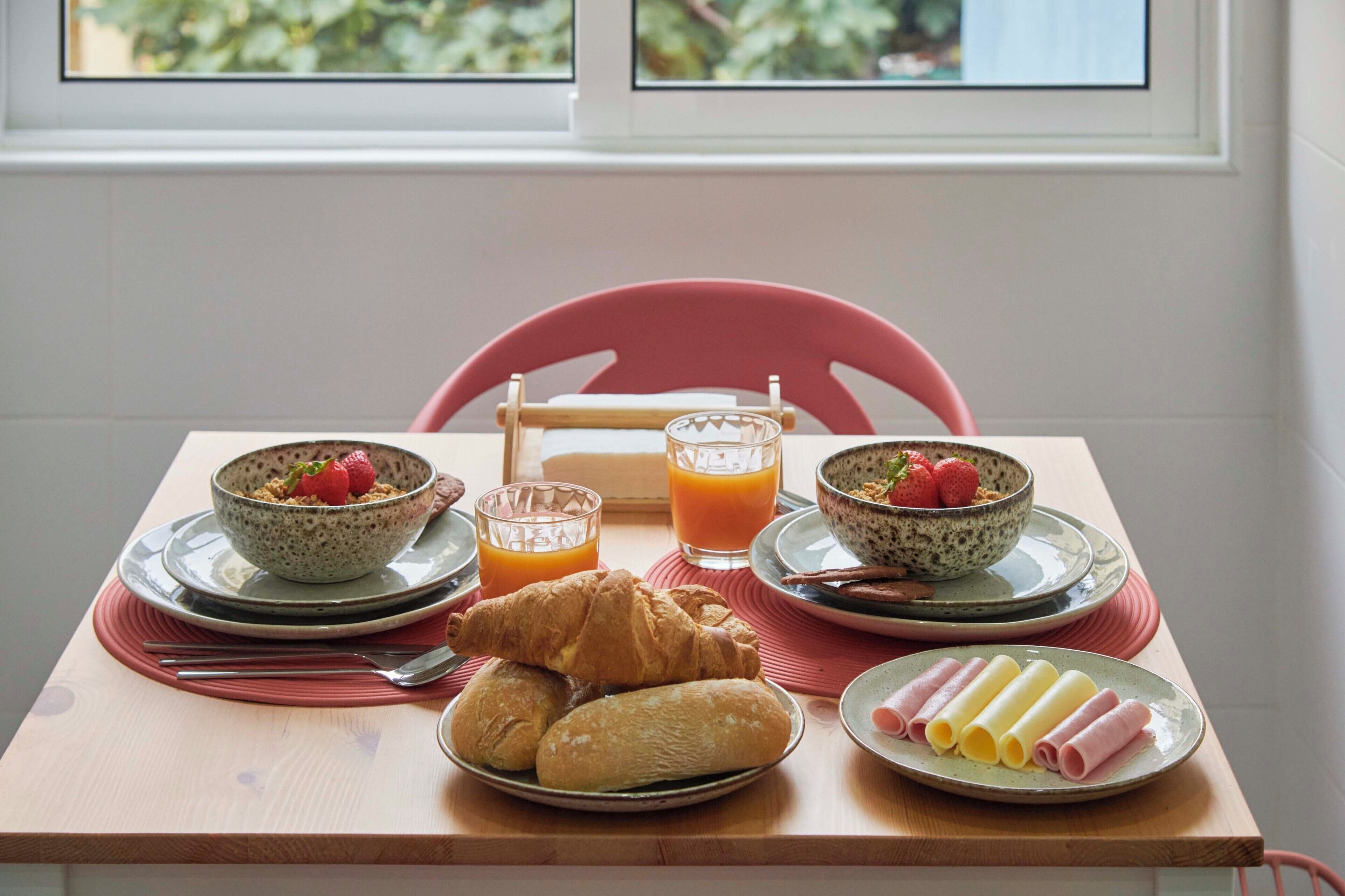 A breakfast table with croissants, ham rolls, and fruit bowls, with a window in the background.