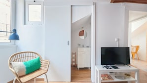 Una habitación con una televisión sobre una mesa blanca, una silla de mimbre con cojín, una puerta blanca y una ventana con persianas.