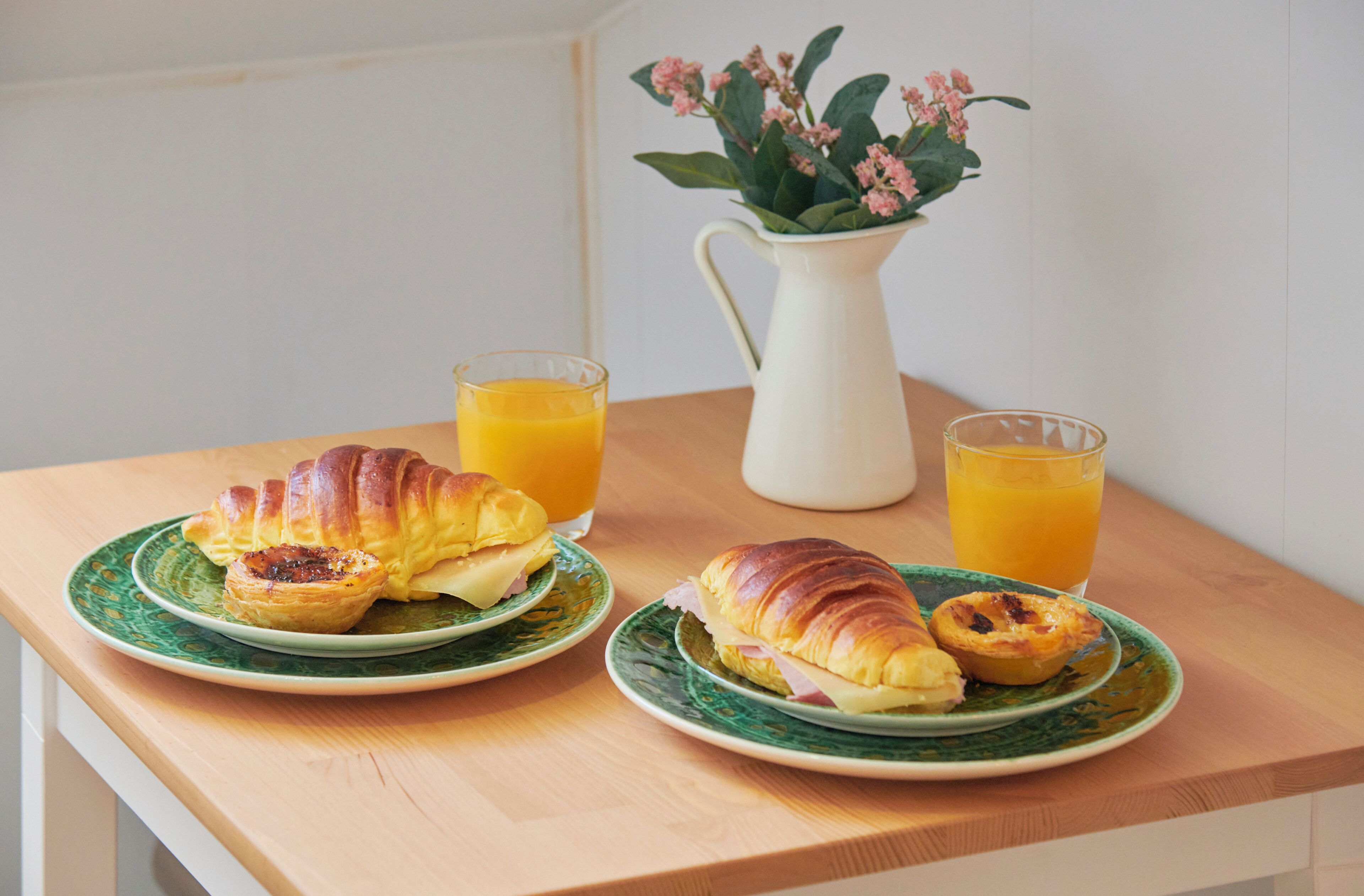 Une table en bois dressée pour le petit-déjeuner, avec des croissants, une carafe et des verres de jus d’orange.