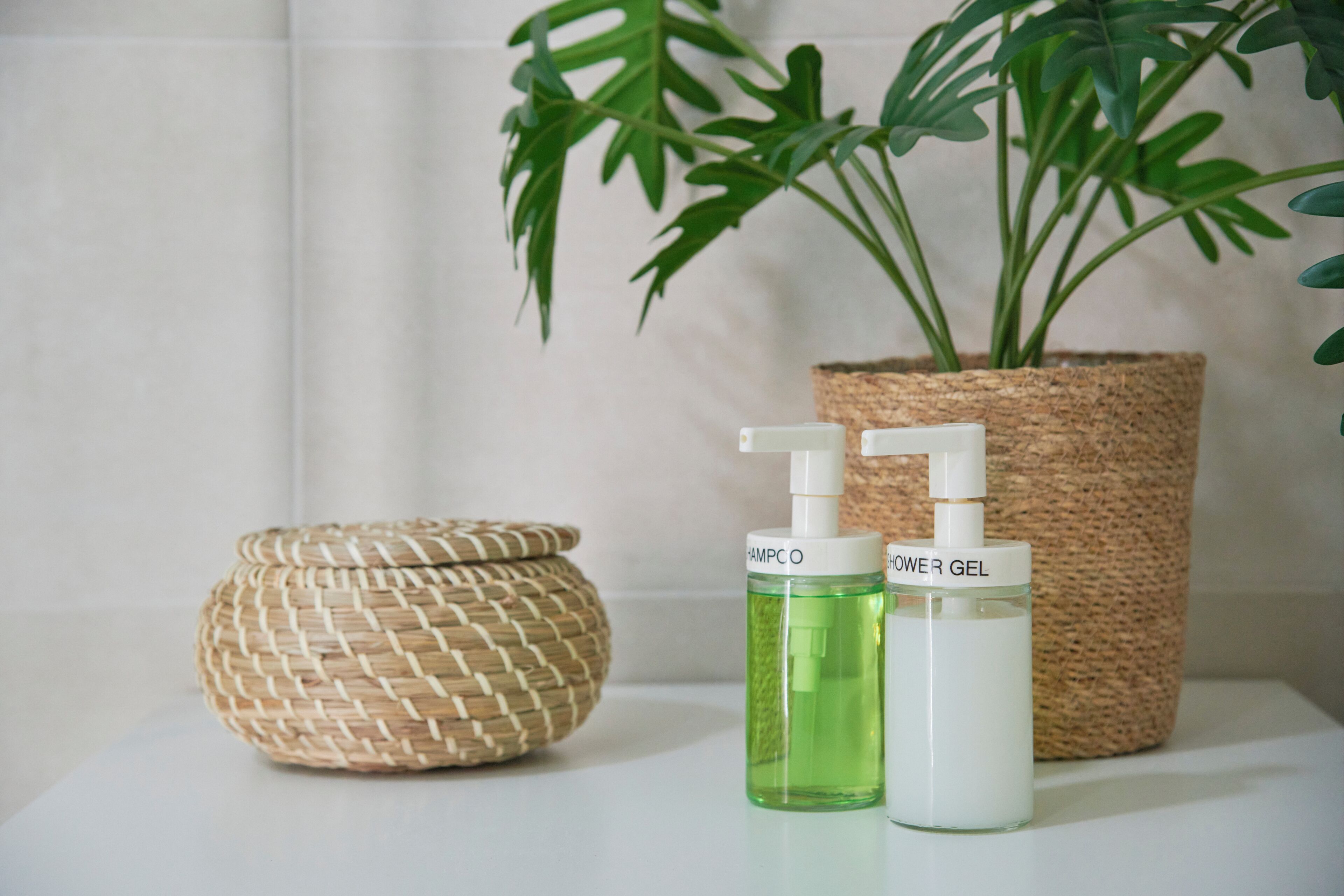 A bathroom with a woven basket, two bottles of shampoo and conditioner, and a potted plant.
