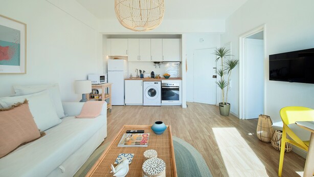A modern living room with a sofa, coffee table, and a dining area with a yellow chair. A kitchen with white cabinets and appliances is visible in the background. - Seaview & Sunset Balcony Charm, By Timecooler (Setubal)