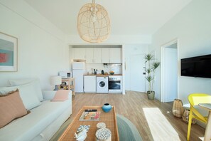 Una sala de estar moderna con un sofá, una mesa de centro y un área de comedor con una silla amarilla. Se ve una cocina con armarios y electrodomésticos blancos al fondo.