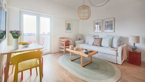 A living room with a sofa, a coffee table, a dining table with chairs, a wicker chair, and a view through a large window. - Seaview & Sunset Balcony Charm, By Timecooler (Setubal)