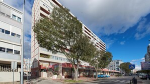A multi-story building with a tree in front, a pedestrian crossing sign, and parked cars. - Seaview & Sunset Balcony Charm, By Timecooler (Setubal)