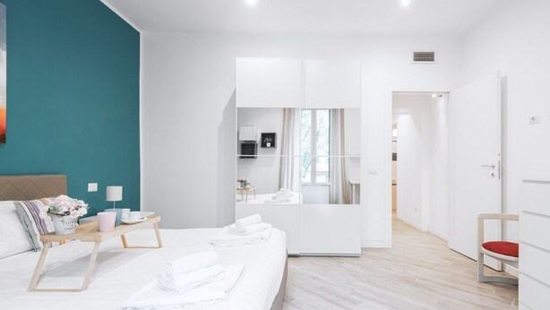 Camera d'albergo moderna con un letto grande, un comodino con una lampada, uno specchio e vista sulla cucina e sul bagno attraverso una porta aperta.