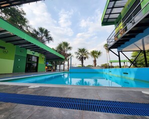 A swimming pool with a modern design, surrounded by a covered area with a green and blue color scheme, and palm trees in the background. - Encanto Llanero by XKP (Villavicencio)