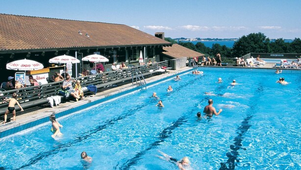 Ein Schwimmbecken mit mehreren Bahnen, Zuschauern und Sonnenschirmen.