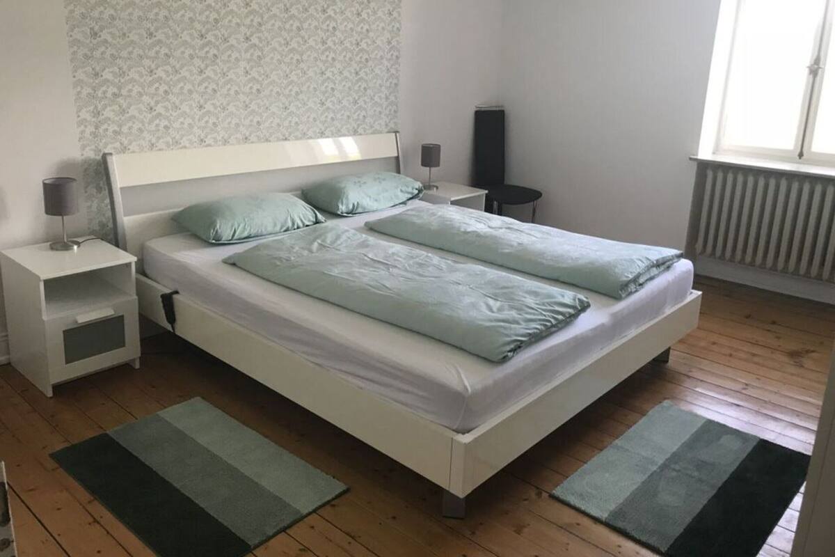 Una cama doble con dos almohadas, una mesita de noche con lámpara, una mesita pequeña y un radiador.
