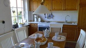 A kitchen with wooden cabinets, a stove, and a sink. There is a dining table set with plates, cups, and a basket of bread.
