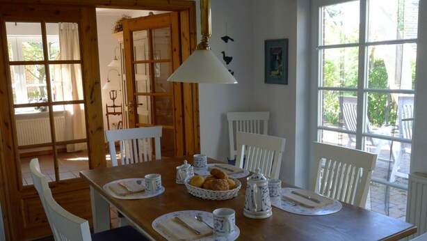 A dining area with a wooden table set for four, white chairs, and a large window providing a view of the outdoors.