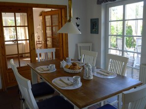 A dining area with a wooden table set for four, white chairs, and a large window providing a view of the outdoors. - Holiday Home Brigitte (St. Peter-Ording)