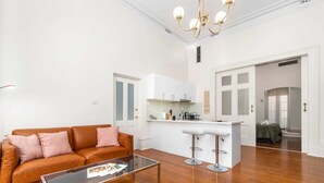 En moderne stue med en brun lædersofa, et glasbord og et køkkenområde med hvide skabe og en barstol.
