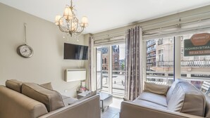 A modern living room with a sofa set, a flat-screen TV, a clock, and a view of the city through large windows. - Cosy Studio Near the Center (Blankenberge)