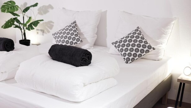 A neatly made bed with white linens and patterned pillows, a potted plant, and a bedside table with a lamp. - Kürzlich Renovierte 3-bett-ferienwohnung mit Garten und Blick auf den Gletscher (Kaprun)