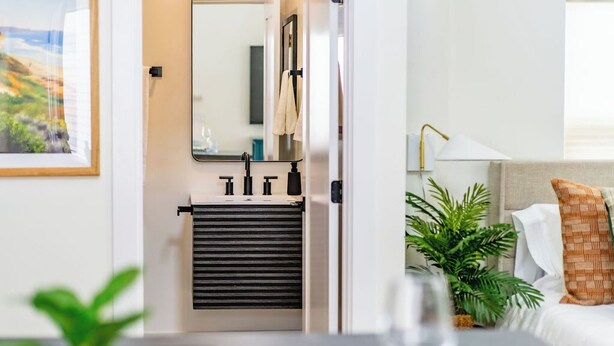 A hotel room with a bed, a bedside table with a glass, a lamp, a potted plant, and a window with a view of greenery. - Swanky Studio in Vibrant Hillcrest (San Diego)