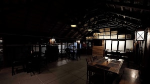 Un intérieur à la lumière tamisée, avec des tables et des chaises en bois, un plafond haut avec des poutres apparentes et une grande fenêtre laissant entrer la lumière naturelle.
