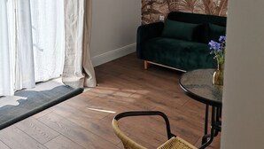 Una sala de estar con un sofá verde, un sillón de mimbre, una mesa redonda de vidrio con un florero y un mural de follaje tropical en la pared.