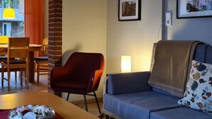 A living room with a sofa, a red chair, a dining table with chairs, and a lamp. There are three framed pictures on the wall.