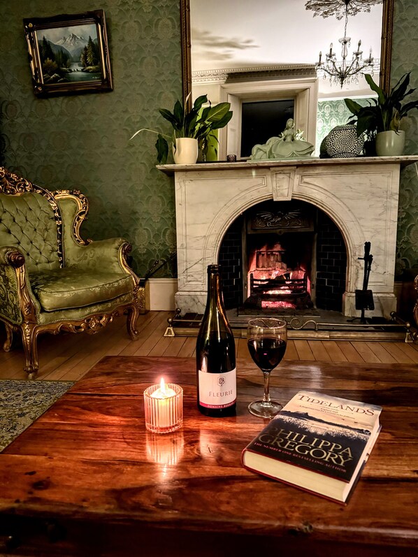 Un cuarto acogedor con un sillón verde, una chimenea, una botella de vino, una copa de vino, una vela encendida y un libro titulado "Tidelands" de Philip Gregory.